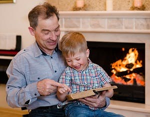 Smiling grandfather and laughing grandchild reading a book in front of a fireplace