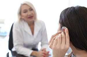 Woman adjusting hearing aid while audiologist looks on