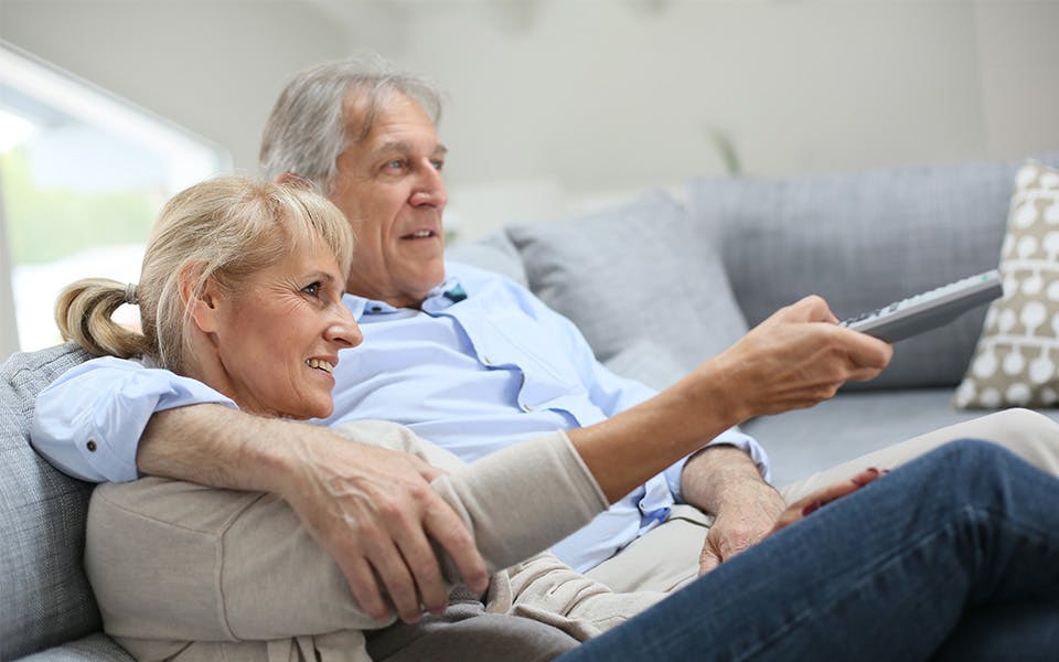 Senior couple sitting on a sofa watching TV with subtitles on.
