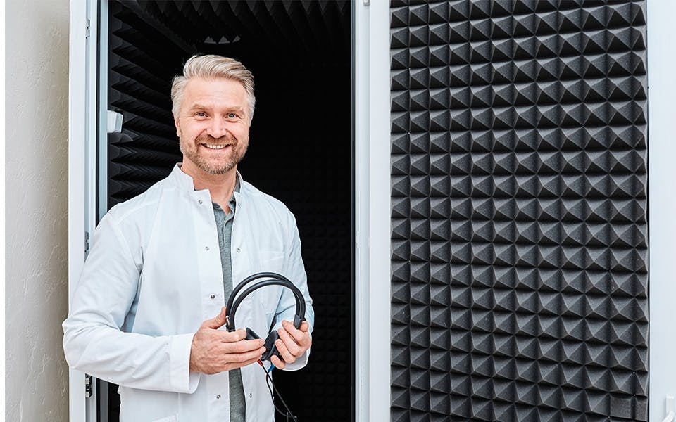 Audiologist standing near soundproof audiometric booth with audiometer headset in hands